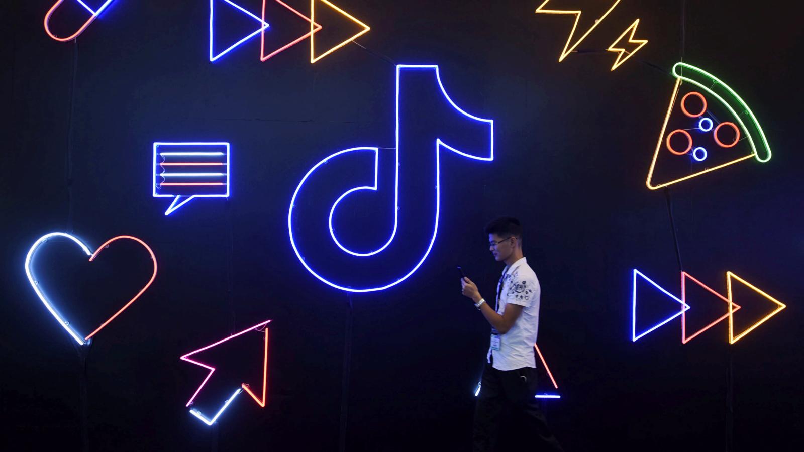 A person standing next to neon signs of heart, pizza, mouse pointer and Tiktok logo