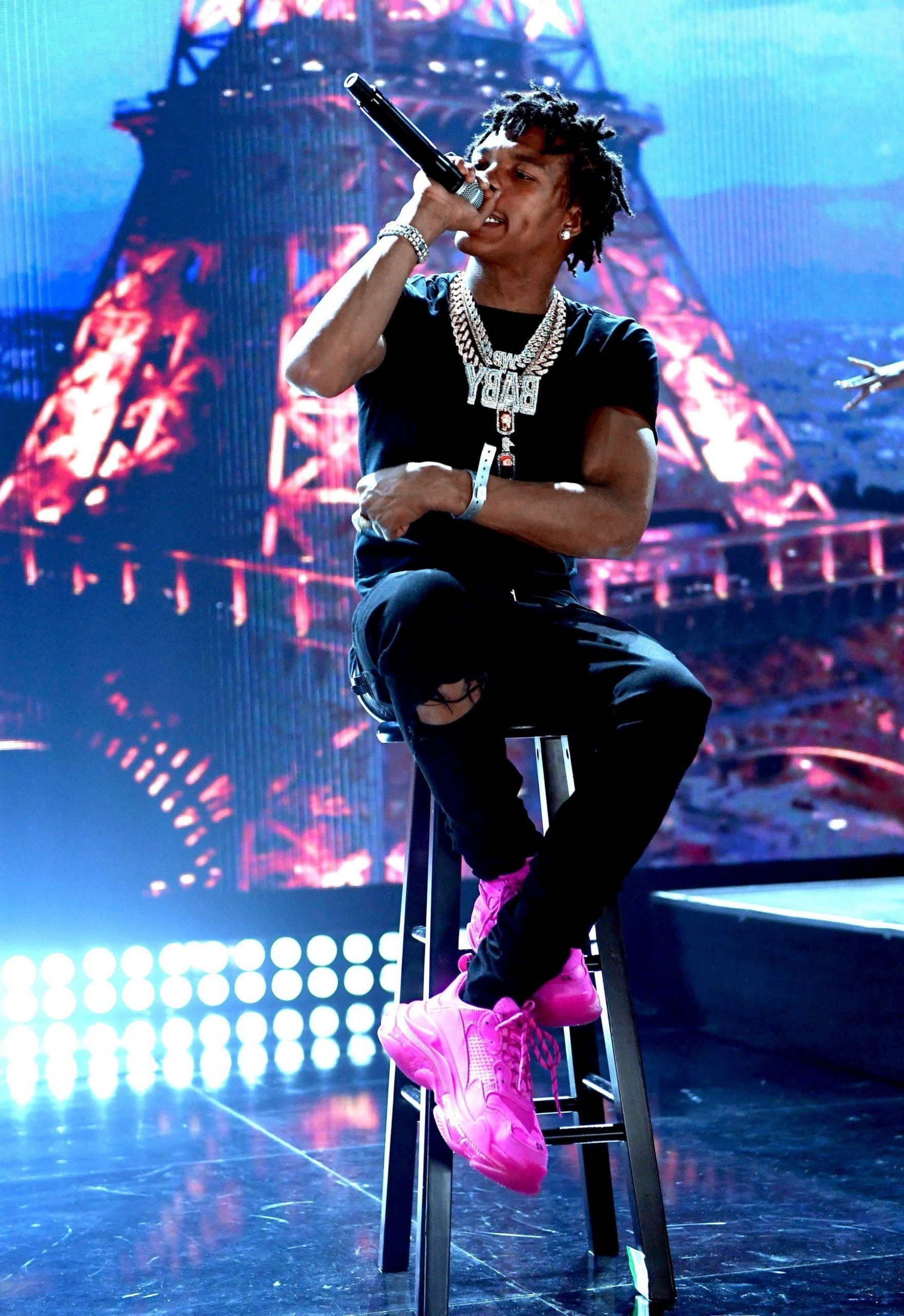 Lil Baby holding Mic onstage while sitting on a stool and wearing pink sneakers