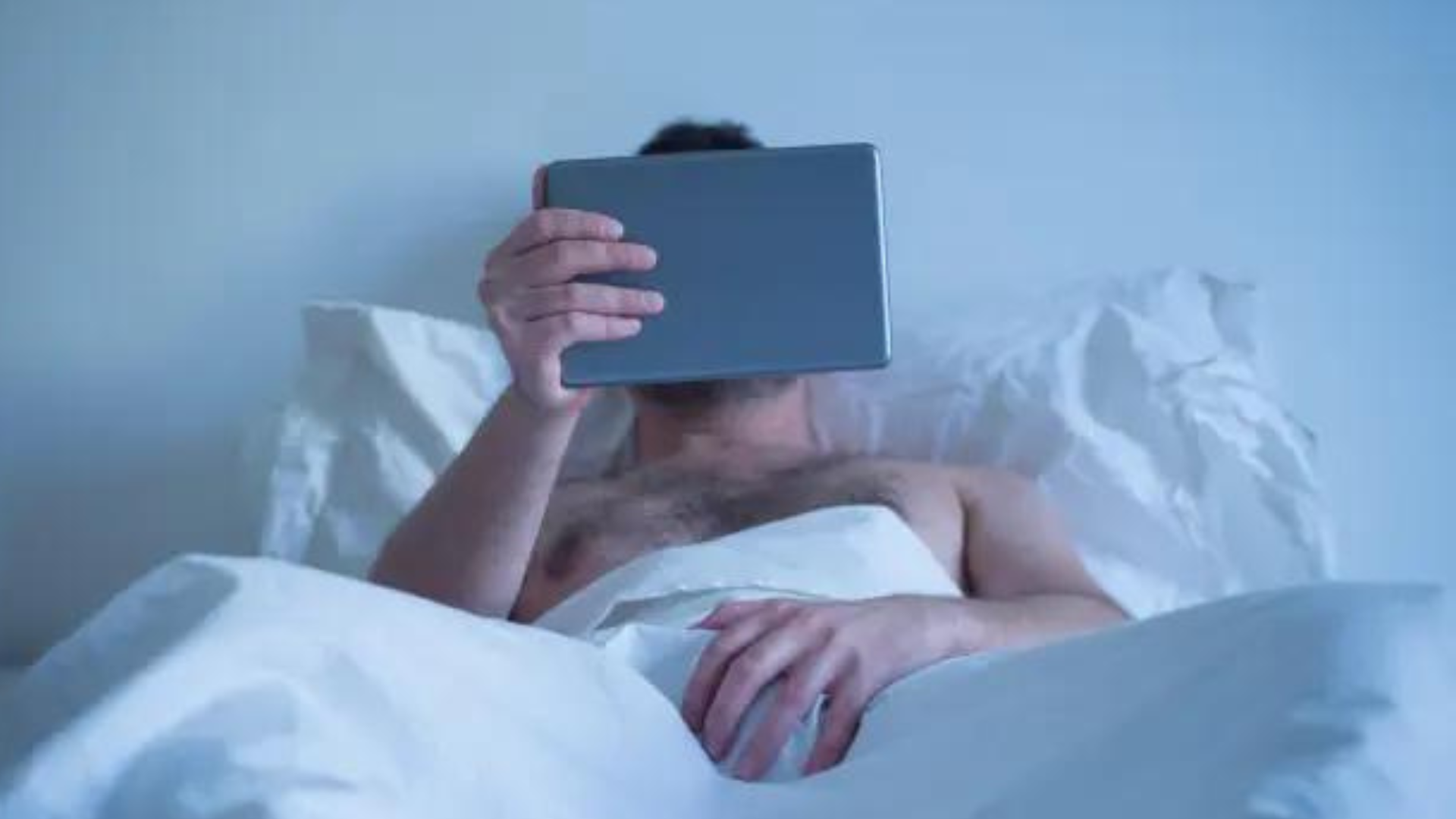 A man lying in bed and watching from his ipad with his hands on his penis